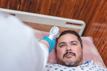 A male patient in his late 30s has his temperature checked with a non-contact scanner while lying on the bed of a ward at a hospital or clinic.