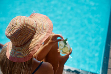 Woman with sun hat drinks summer cocktail by pool.