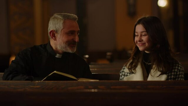 A priest sits in a church with a bible in his hands talking to a parishioner. A Catholic priest communicates with a smiling brunette woman after the Sunday service.