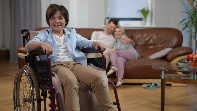 Happy smiling boy in wheelchair looking back at hugging parents sitting on couch and looking at camera. Portrait of confident Caucasian son posing in living room at home with father and mother
