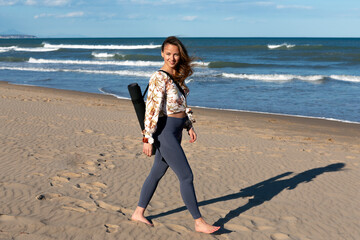 young woman walking barefoot on the beach with mat slung over her shoulder