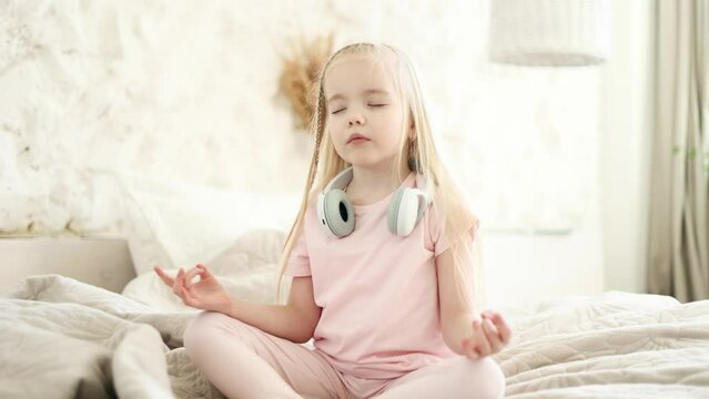 Calm relaxed blond little girl child keeps hands in yoga gesture meditating and breathes slowly while make lotos asanas on bed in the morning before school at home No stress happy people concept