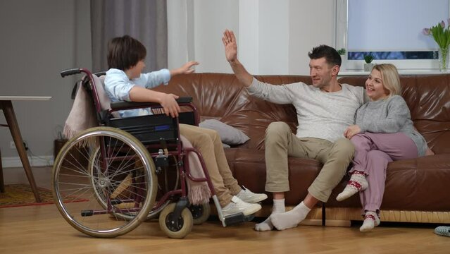 Wide shot couple of parents talking sitting on sofa as son in wheelchair entering rolling gesturing high-five with father. Positive confident Caucasian family with disability indoors