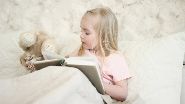 Adorable cute little blond girl child kid reading book fairy tale with her soft bear toy friend while laying in bed in the morning at home Happy childhood concept