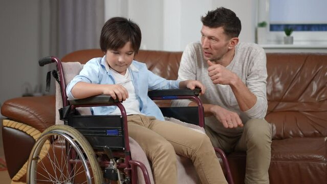 Curious cute boy in wheelchair talking with man sitting on sofa in living room. Portrait of positive confident Caucasian son and father enjoying leisure together at home indoors