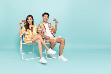 Happy Asian couple sitting on beach chair and showing credit card isolated on green background,...