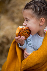 A girl wrapped in a yellow cloth eats a cinnamon bun and nuts against the background of straw bales