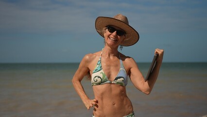 Pretty mature senior woman in hat sunglasses and bikini holding laptop at the beach showing freedom...