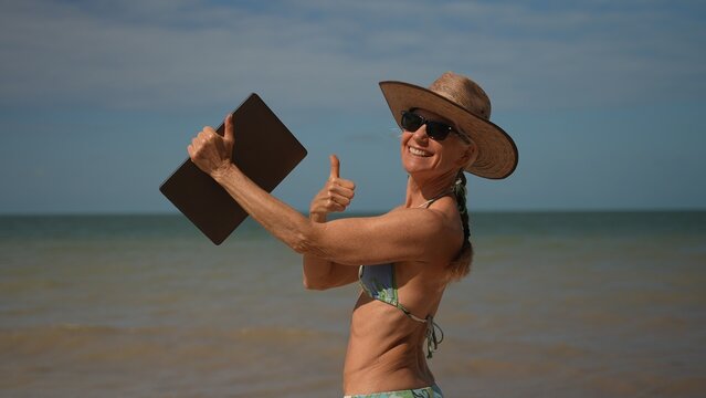 Pretty happy smiling mature senior woman in hat sunglasses and bikini holding laptop at the beach giving thumbs up as digital nomad in tropical place.