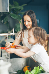 Mother and two-year-old daughter wash vegetables in the kitchen sink. Cooking and health care. Side view.