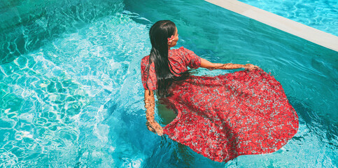 Summer dress fashion model woman swimming in pool wearing long floral red maxi dress clothing. Creative artistic fashion photo shoot at luxury resort infinity pool