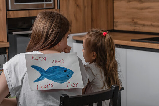 Mom and daughter are sitting in the kitchen, on the back of a young woman is a paper fish with the inscription April Fool's Day. Funny family jokes, celebration.