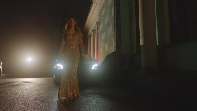 View of luxurious beautiful blond woman in golden elegant dress walking slowly in the night at car lights, black tie. Theater, museum, historical building exterior. Concert, show. Slo mo HQ 4k footage