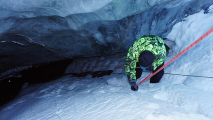 A climber descends by a safety rope into an ice cave in a glacier. The gray-blue color of ice. White snow. The guy has a bright jacket. Red rope. A dangerous descent into the unknown.