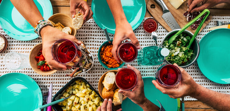 above view of table with group of friends people celebrating and toasting with red wine glasses and eating food all together having fun. Lunch time and friendship. Family celebrate clinking drinking