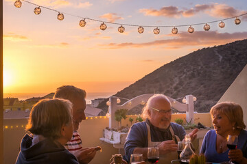 Happy group of senior people enjoy dinner time with sunset and town in background. Mature elderly...