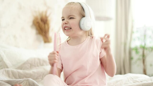 Funny expressive happy little girl child in headphones sitting on bed and singing in hairbrush microphone at home Cute blond kid enjoying free time in the light living room alone