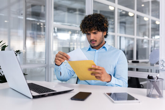 Young businessman at work inside office received mail envelope notification message with bad news, hispanic man sad reading letter document sitting at table with laptop.