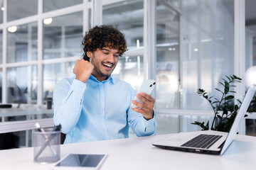 Young successful businessman inside the office at work uses a laptop hispanic man holds a smartphone in his hands, the man holds his hand up a gesture of success and triumph, celebrating victory.