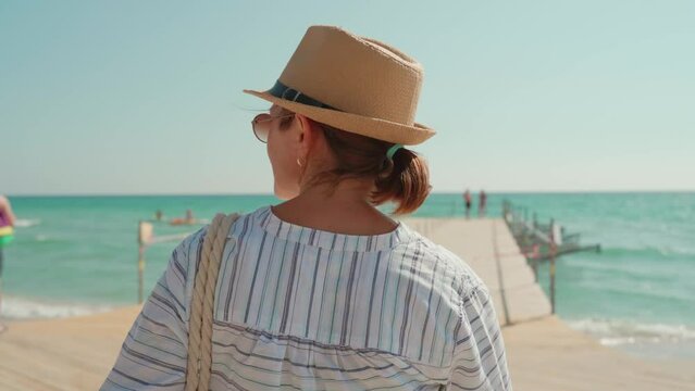 a woman in a hat and a striped shirt walks along the pier into the sea. back view