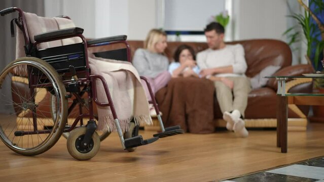 Wheelchair on the left with blurred ill son mother and father at background on couch. Wide shot living room with positive Caucasian unrecognizable family resting at home. Health care and disability