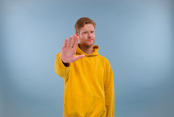 Serious man wearing a yellow hoodie shows a hand gesture STOP and looks at the camera with a dissatisfied face, isolated on a blue background.