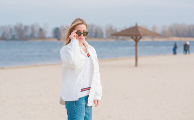 Woman in white jacket, sunglasses and blue jeans at the beach, new collection, fashionable trend