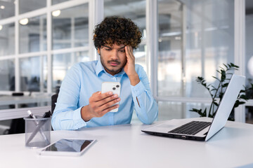 Businessman inside the office at the workplace is not satisfied with the work of the phone, the man is upset holding a smartphone in his hands, hispanic man is sitting at the table with a laptop.