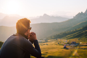 Person mit Sonnenbrille sitzt in den Bergen bei Sonnenuntergang und genießt die Natur  - obrazy, fototapety, plakaty