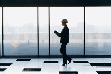 Fototapeta na wymiar the silhouette of a girl in a white construction helmet walks near the panoramic window. girl architect designer