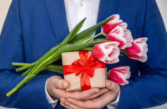 man holding one tulip flower in hand or in jacket on palm.male in office suit with gift box and tulips bouquet faceless woman mother international day holiday march 8th.spring is coming birthday
