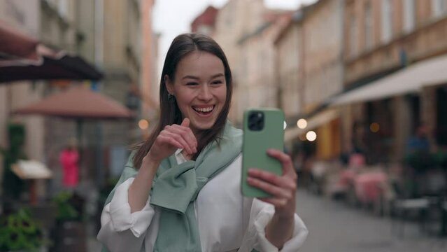 Attractive Deaf-mute woman saying phrase Hello, how are you with sign language during video call on modern smartphone. Girl walking on city street and enjoying distance communication.