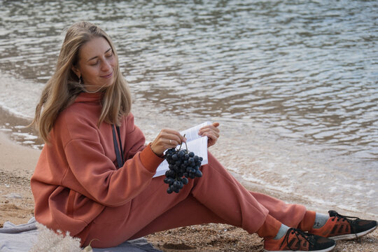 A girl in an orange tracksuit, hoodie and pants sits on the beach and enjoys. Sits by the sea and eats fresh bread, fruits, grapes and reads a book. High quality photo