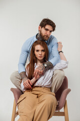 Fashionable beautiful couple sits and poses in a chair on a white background in the studio. Beautiful redhead model woman and brutal hipster handsome man with tattoos