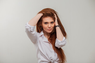 Fashionable beautiful redhead model girl with long red hair in stylish white elegant clothes poses on a grey background in the studio