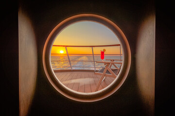 Pont promenade d'un navire de croisière en navigation avec un transat et un cocktail rouge au coucher de soleil à travers un hublot.	