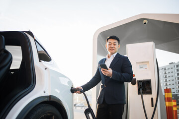 Well-dressed male entrepreneur checking battery on modern smartphone while charging luxury electric car at outdoors station. Alternative automobile and ecology protection concept.