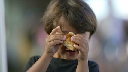 Portrait of child eating piece of bread croissant. Close up face of one small boy kid eating carb food