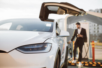 Blurred background of indian man in formal wear restoring battery of his electric car at EV charging station. Focus on white shiny luxury auto. Eco friendly transport.