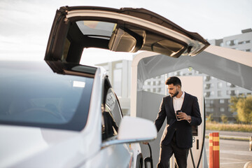 Attractive bearded man in business suit plugging in power cord to electric car while holding coffee cup. Stylish indian male having stop on EV station for charging eco friendly vehicle.