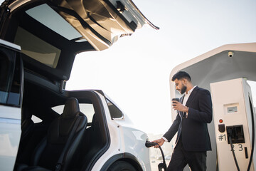 Side view of handsome arabian man in formal outfit holding coffee-to-go in one hand while inserting plug into luxury electric car. Charging process of modern eco transport.