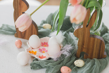 Happy Easter! Beautiful tulips, natural eggs and bunny decoration on modern table. Stylish farmhouse easter decor. Handmade egg holder, pink tulips bouquet and wooden bunnies