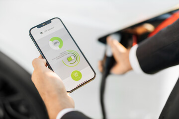 Close up of businessman charging electric car on station and checking process status on modern smartphone. Focus on mobile screen with info from auto app.