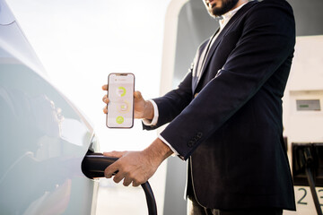 Crop of man in black suit plugging power cord and showing mobile screen with app of charging status...