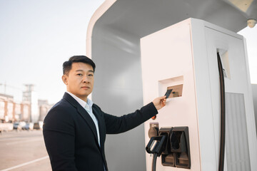 Portrait of handsome man in stylish black suit using credit card while paying for charging electric car at outdoors station. Modern technology in automotive industry and contactless purchase.