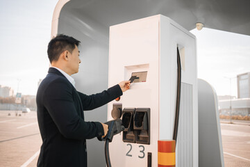 Good-looking asian man touching monitor sensor with credit card while recharging battery of electric car. Successful businessman using cashless system for paying at EV station.