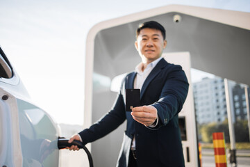 Positive asian man in stylish black suit charging white electric car and showing black credit card outdoors. Cashless payment for energy at EV station. - obrazy, fototapety, plakaty