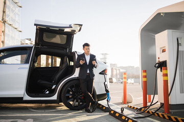 Asian businessman with credit card and modern smartphone in hands paying for energy while charging luxury electric car at urban station. Transport and technology concept.