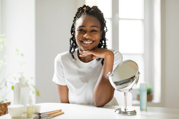Portrait of beautiful black woman looking and smiling at camera, touching face, sitting at dressing table at home