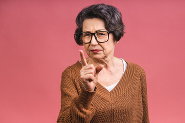 Portrait of angry grey haired old strict senior woman wearing glasses pointing up threatening with finger. Grandmother isolated over pink background. - 578469244